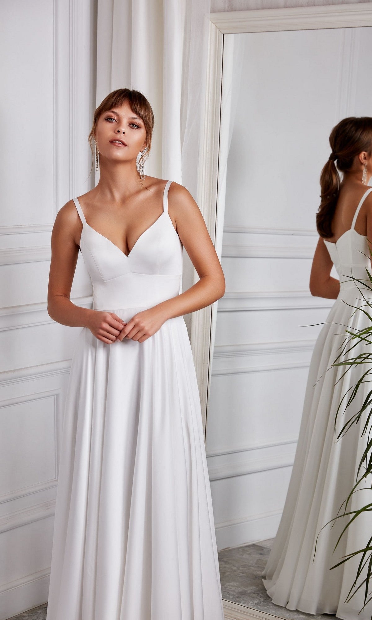 A woman stands indoors near a mirror, hands gently clasped, wearing the Aerie Couture 2003 Long White Dress in Diamond White with a V-neckline and an elegant A-line skirt.